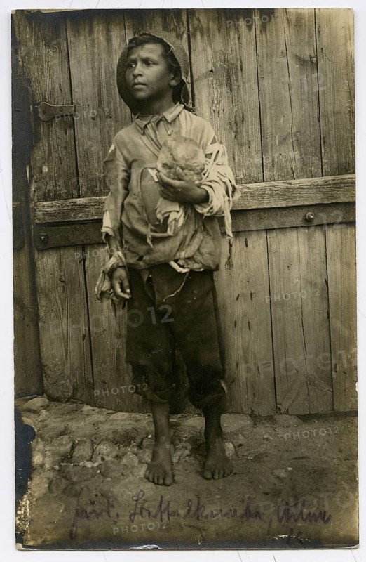 Jewish beggar, boy, Vilnius, Lithuania, poverty, hunger, orphan, boy in tattered clothes holding bread and standing barefoot in front of a wooden wall