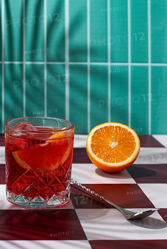 A drink with ice and orange slices is placed in a glass on a checkered table. A cut orange sits beside the glass. Shadows from nearby plants add a touch of detail