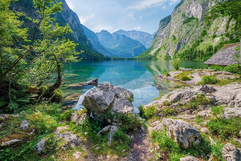 Obersee mountain lake Alps Bavaria Germany with a rocky path, clear turquoise water, alpine cliffs, and sunlit vegetation along the peaceful lakeshore