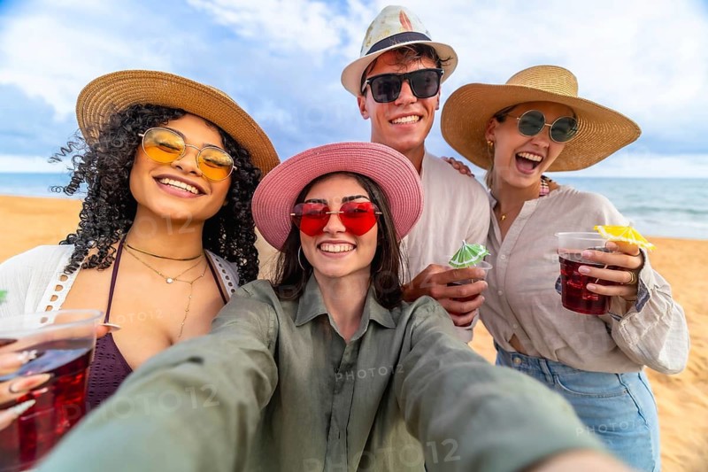 Group of diverse young happy friends laughing and smiling while taking a selfie on a sandy beach, holding refreshing cocktails during tropical summer holiday