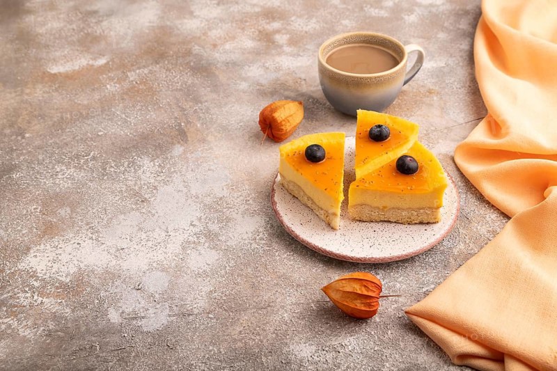 Cheesecake with mango cream, cup of coffee on brown concrete background with orange textile, copy space, side view