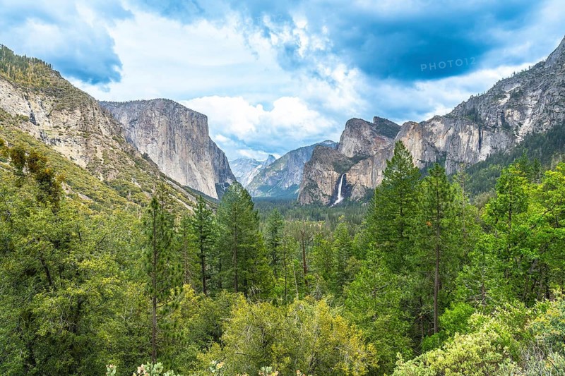 Yosemite valley panorama presenting el capitan, bridalveil fall, and half dome nestled among lush green forests and towering granite cliffs under a vibrant blue sky with dramatic clouds