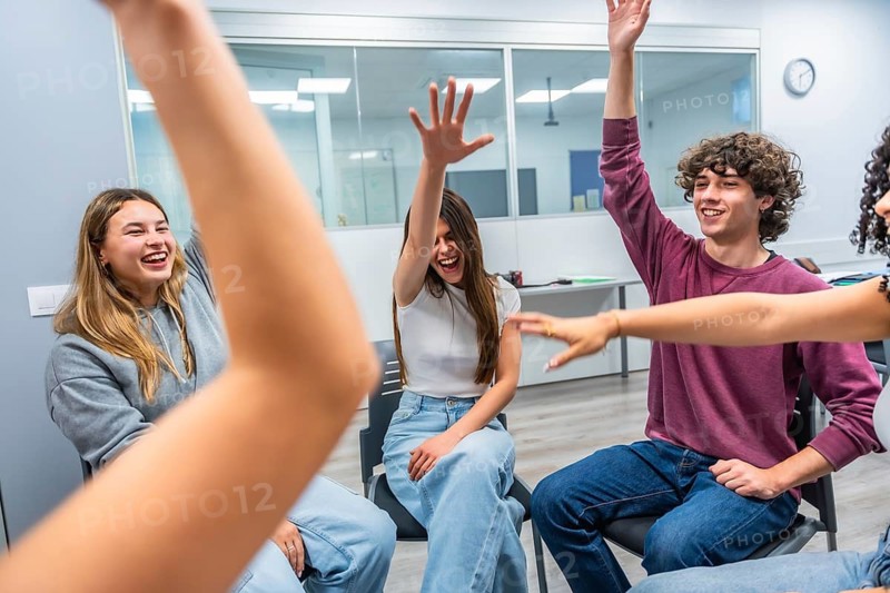 Group of happy, young, diverse students actively participating in a modern classroom setting, raising hands and smiling, showing collaboration and engagement during an educational activity