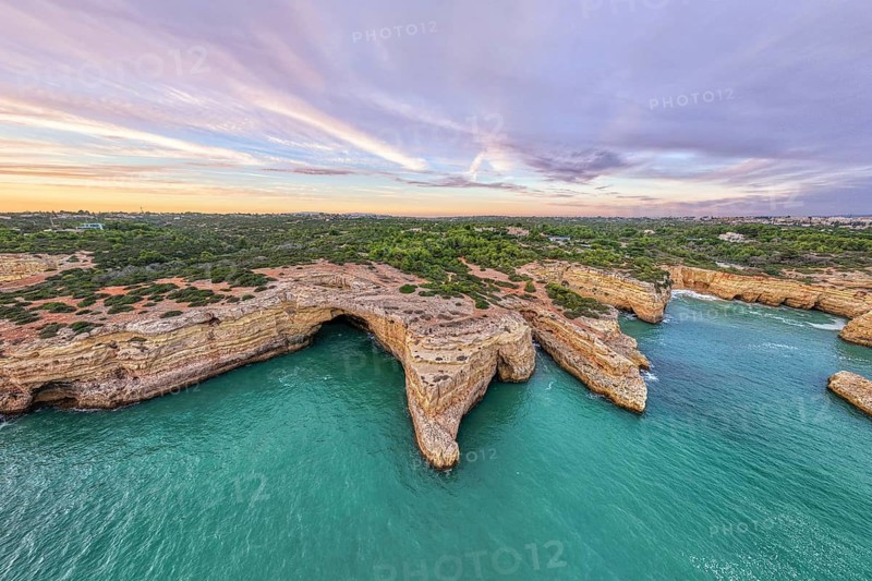 Drone sunset view of Arco de Albandeira at Praia de Albandeira in Algarve, Portugal, with limestone cliffs, Atlantic water, sea arch, and soft pink evening clouds above the coast