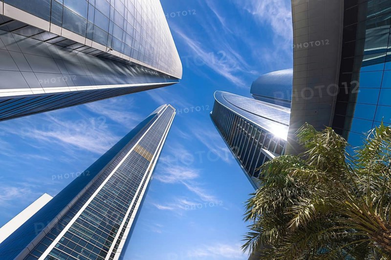 A dramatic low-angle view looking straight up at the modern glass Etihad Towers skyscrapers against a bright blue sky in Abu Dhabi, United Arab Emirates