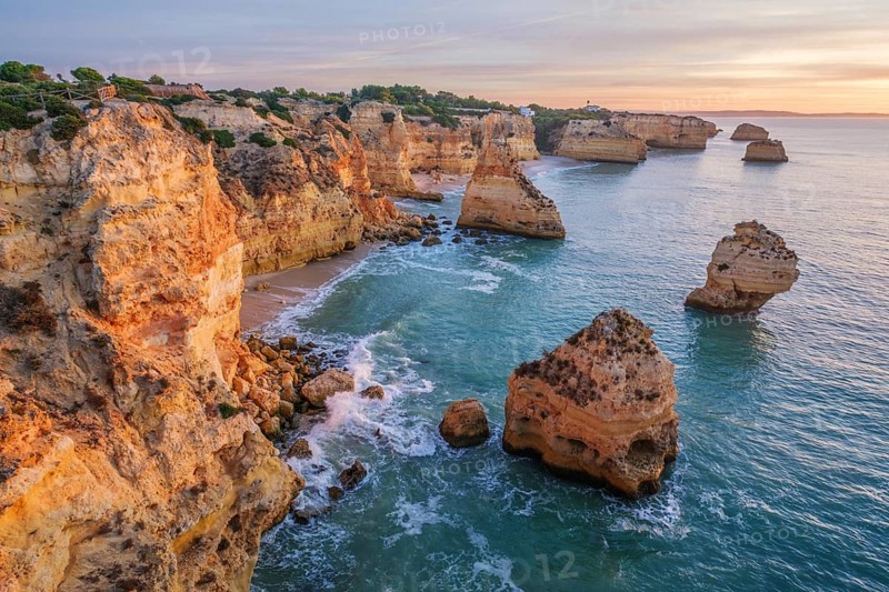 Sunrise view of Praia da Marinha beach in Algarve, Portugal, with a limestone sea arch, Atlantic water, warm sky, and rugged coastal cliffs