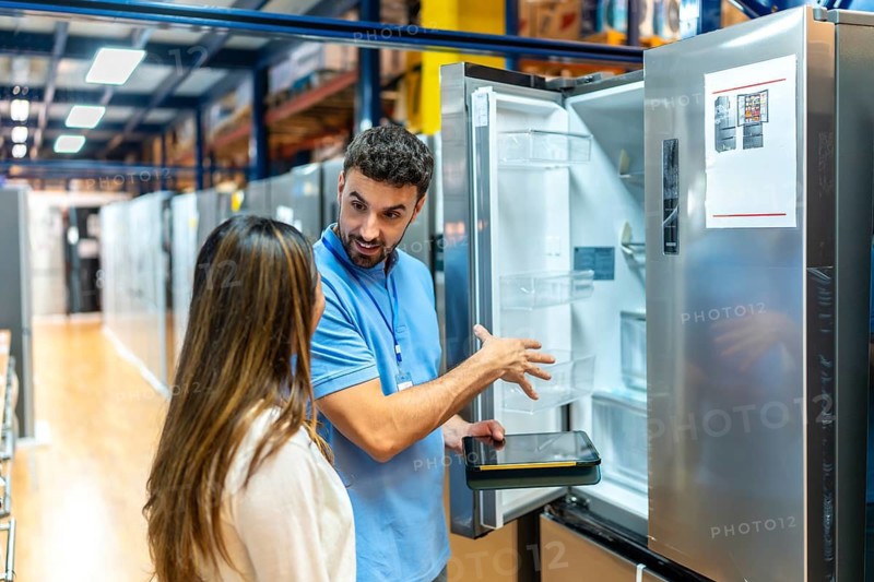 Salesman helping a female customer choose a new refrigerator, pointing out features inside the open door while holding a tablet in a retail store setting