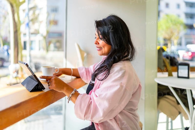 Mature woman sitting at a counter in a bright cafe, smiling while interacting with a digital tablet and enjoying a cup of hot coffee, embracing modern lifestyle and technology