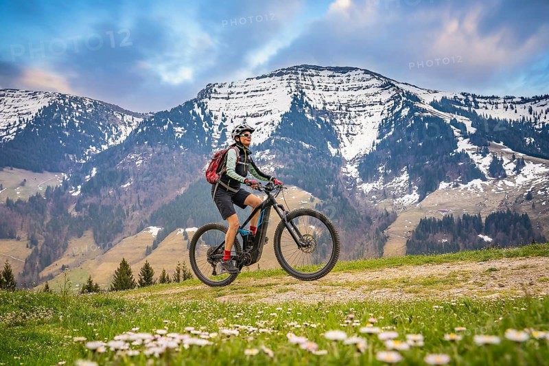Pretty mid age woman riding her electric mountain bike in early springtime in the Allgau mountains, below the spectacular snow capped mount Hochgrat, Nagelfluh chain, Oberstaufen-Steibis, Bavaria, Germany