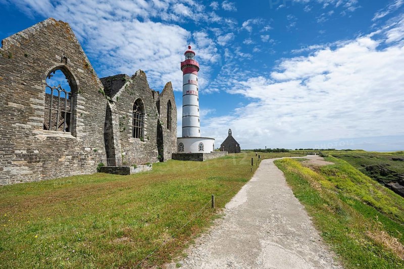 View of the 37 meter high, red-and-white striped lighthouse Phare de Saint-Mathieu, which rises majestically amidst the historic ruins of an old medieval former Benedictine abbey, Abbey Saint-Mathieu de Fine-Terre. A paved footpath leads through a green coastal landscape under blue summer skies with white clouds directly to the historic landmark. Pointe Saint-Mathieu, Plougonvelin, Arrondissement of Brest, Finistère Department, Brittany Region, France