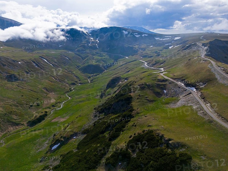 Road runs through green mountain hills in a vast valley under cloudy sky, aerial view, near Rânca, Ranca, Transalpina High Road, Drum Na?ional 67C, National Road, Transylvania Alps, Southern Carpathians, Romania