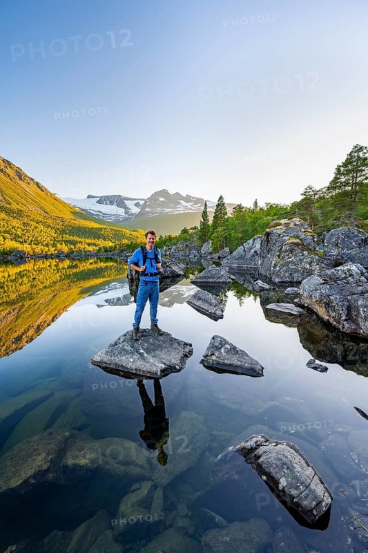 Young man standing on a rock in water, enjoying the landscape, picturesque mountain scenery in the Innerdalen high valley, mountain peaks reflected in Lake Innerdalsvatna, in the evening light at sunset, Trollheimen Mountain Area, Sunndal, Møre og Romsdal, Norway