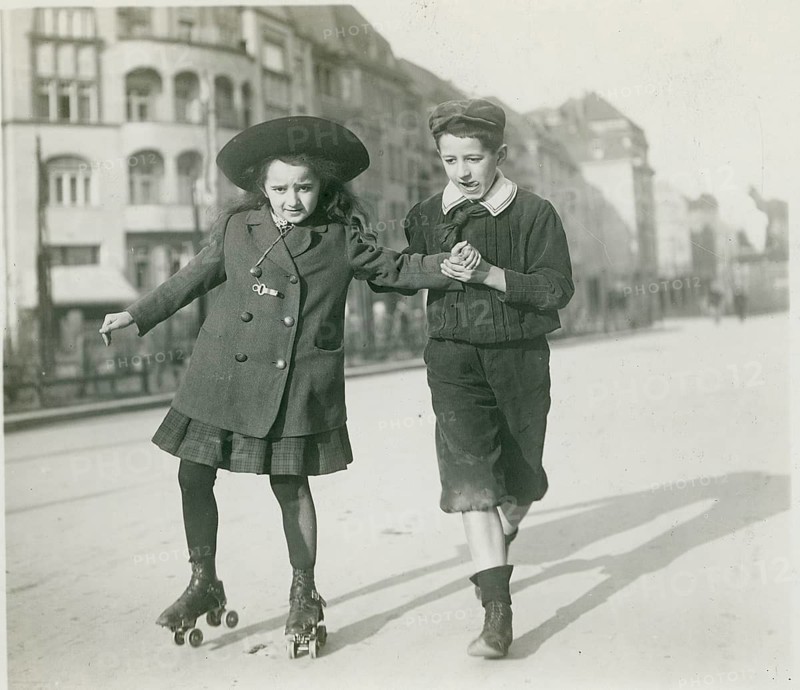 Berlin, roller skating, bourgeoisie, leisure, street, two children in old-fashioned clothes, the girl wearing roller skates while the boy supports them. In the background, an urban backdrop, Philipp Kester, around 1906