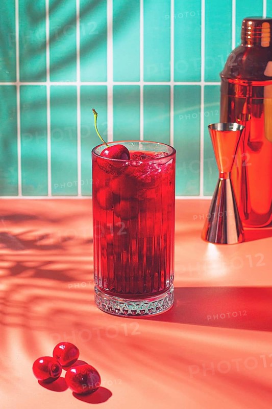 A bright drink made with cherry sits on a table next to some fresh cherries. The background shows a colorful wall and a cocktail shaker. Sunlight casts shadows
