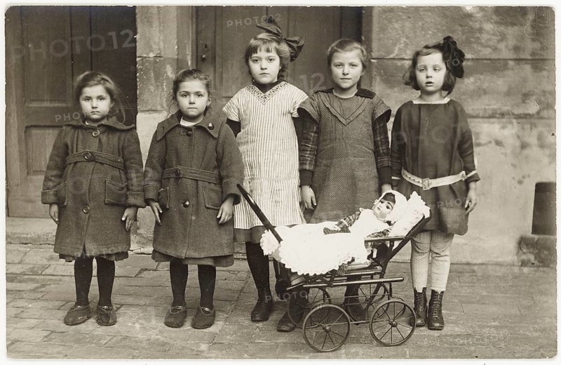 Children, backyard, twenties, big city, doll stroller, twins, five girls standing together, one holding a trolley with a doll. The style looks old-fashioned, around 1920
