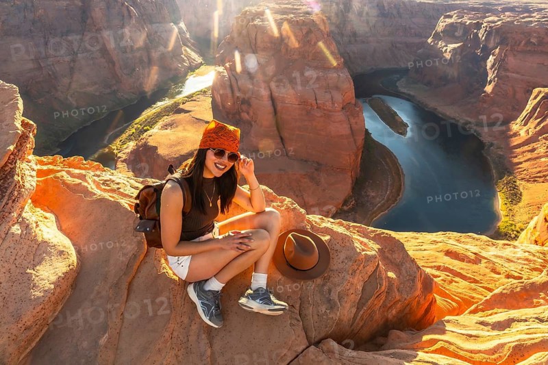 Young woman smiling while sitting on a cliff edge overlooking the winding colorado river at horseshoe bend, enjoying a scenic travel destination with clear blue sky