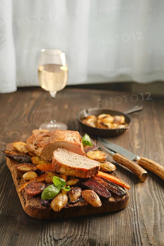 Dinner features roasted meat and vegetables served on a wooden board. A glass of white wine is placed nearby. This setting is indoors, with soft natural light coming through a window