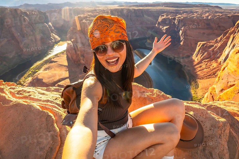 Young happy woman sitting on a cliff taking a selfie with horseshoe bend and the colorado river in the background during a sunny sunset hike demonstrating travel and freedom