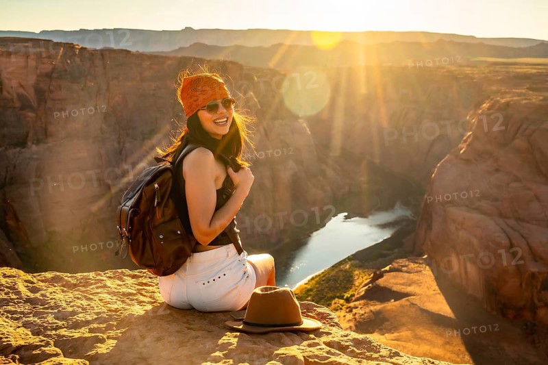 Young woman traveler with backpack and bandana smiling, sitting on a rocky cliff overlooking the winding colorado river within the majestic canyon landscape at horseshoe bend during a golden sunset