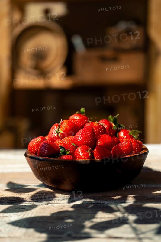 Bowl filled with ripe strawberries on wooden table under bright sunlight. Hard shadows fall around bowl, creating warm atmosphere. Background shows wooden decor giving rustic feel