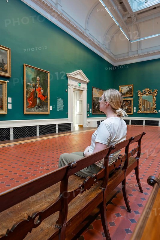An art viewer sits on a wooden bench in a gallery surrounded by paintings on green walls, National Gallery of Ireland, Dublin, Ireland