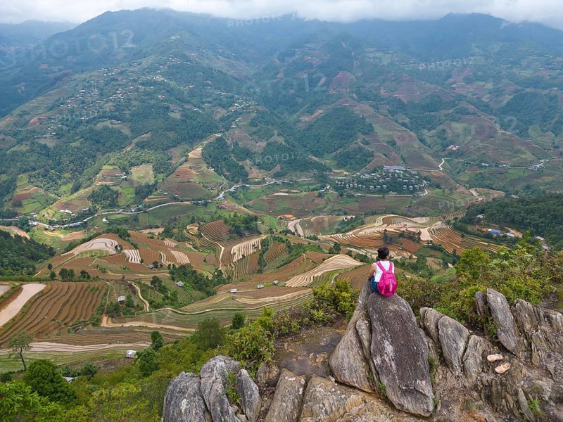 Aerial view of a woman with a pink rucksack sitting on The Dinosaur Backbone (S?ng Lung Kh?ng Long), De Xu Phinh Commune, looking out over rice terraces during the planting of the new, fresh-green rice seedlings at Mù Cang Ch?i, Mu Cang Chai District, Lào Cai Province, Northern Vietnam