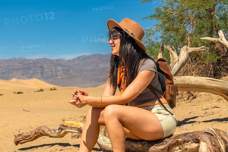 Woman wearing a hat and backpack sitting on dry wood and looking at sand dunes with mountains in the background, enjoying the silence and natural beauty of death valley national park