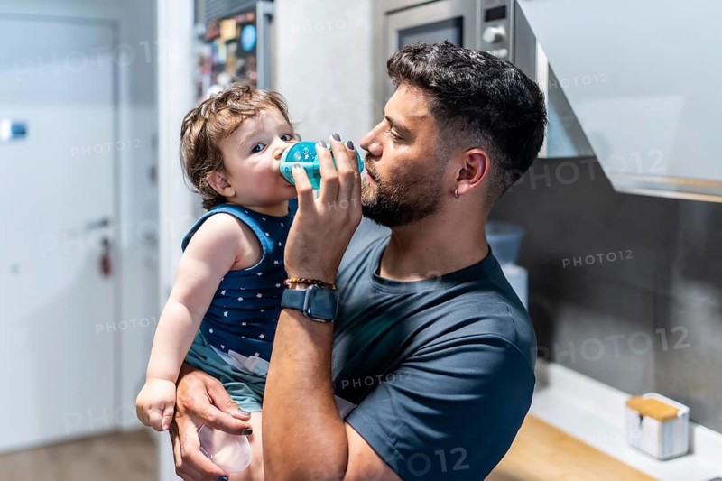 Father gently helps toddler son drink from a sippy cup in the kitchen, capturing a tender moment of hydration, care and bonding between dad and child at home