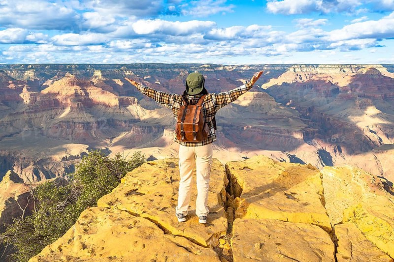 Person with outstretched arms expressing freedom and adventure while standing on a rocky outlook, admiring the vast landscape of the grand canyon national park under a bright sky
