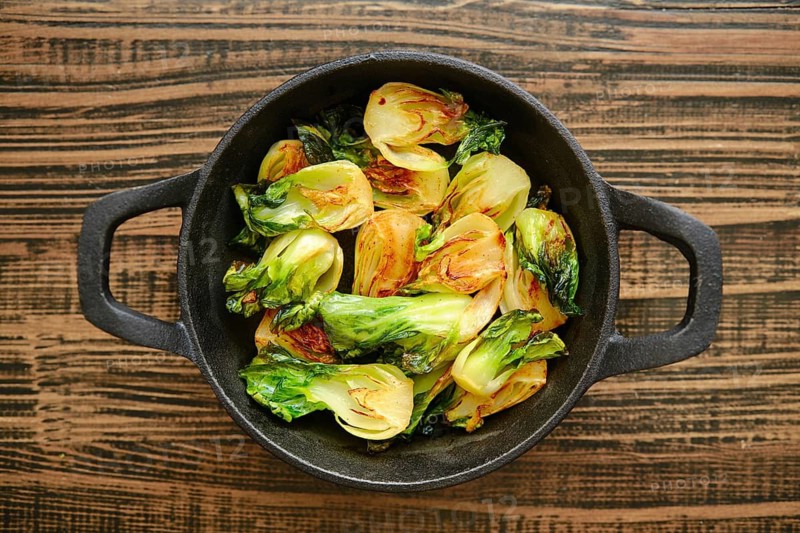 Bok choy is being cooked in a cast iron skillet. The greens are heated and show signs of roasting on a wooden surface. The kitchen setting gives a simple feel to the cooking process