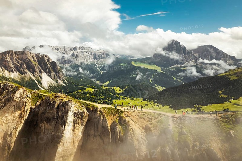 Aerial panorama tourist walk hike Seceda ridge with Odle peaks rise over clouds with steep limestone cliffs green alpine slopes winding hiking trail. Panoramic Dolomites landscape. South Tyrol Italy