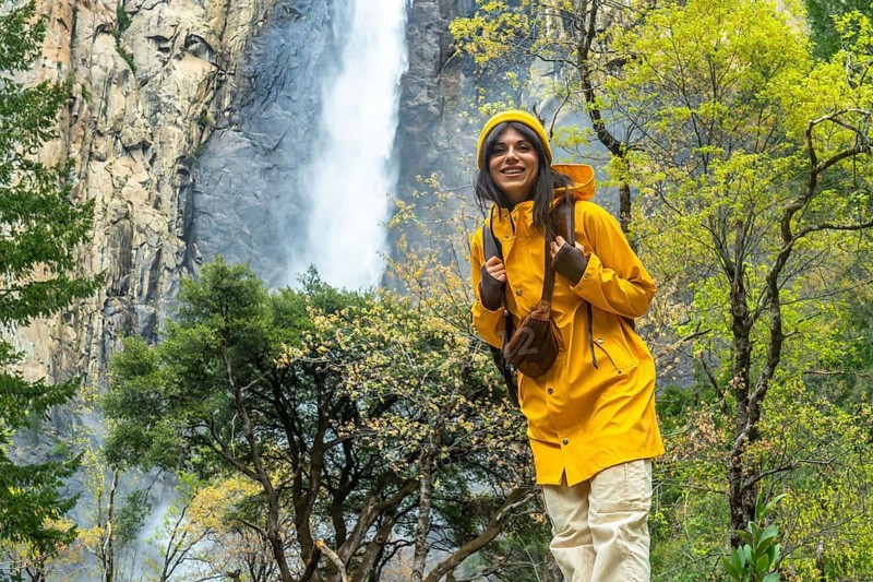 Smiling woman wearing a yellow raincoat and hat, carrying a backpack, standing on a trail near bridalveil fall in yosemite national park, surrounded by spring foliage