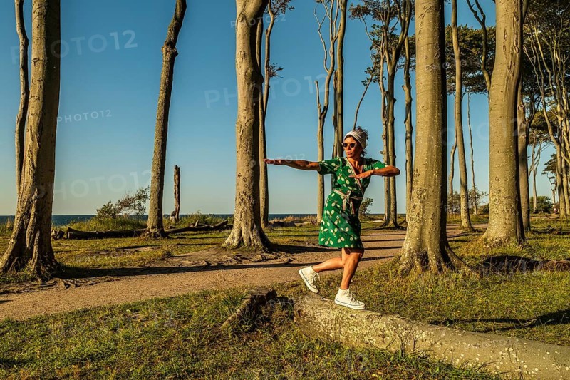 A woman (54) in a green tennis dress and white trainers is surfing on a tree, Ghost Forest, Nienhagen, Germany