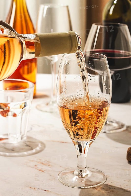 A person is pouring wine from a bottle into a clear glass. There are other drinks on the table, including glasses of red wine and a bottle of white wine
