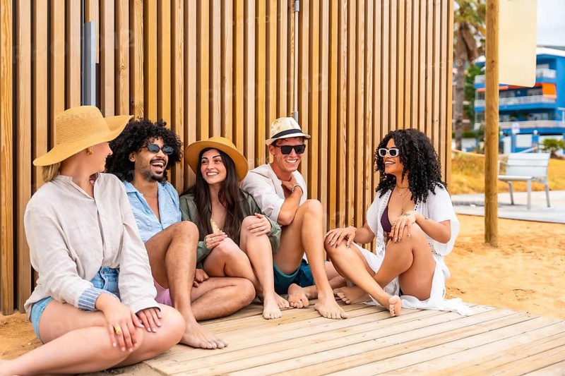 Group of diverse friends wearing casual summer clothing and sunglasses, sitting on a wooden boardwalk near the beach, laughing and enjoying their leisure time during summer holidays