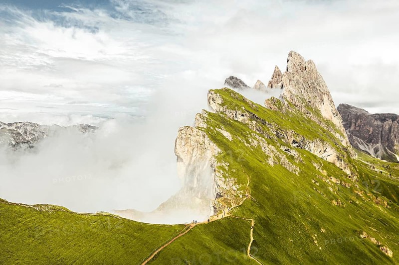 Aerial panorama Seceda ridge Odle peaks rise above clouds after rain with steep limestone cliffs green alpine slopes winding hiking trail panoramic Dolomites landscape South Tyrol Italy