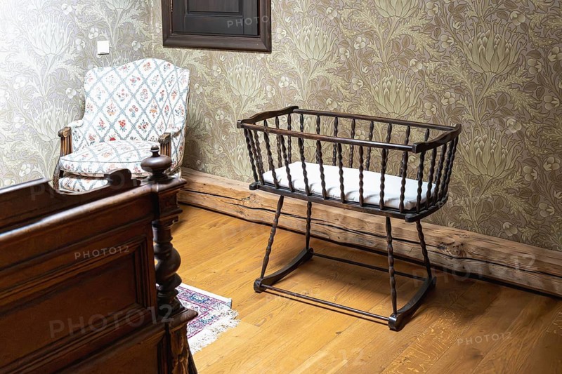 A vintage style room features a dark wooden baby cradle with spindled sides and a white mattress. Nearby stands an upholstered armchair with patterned fabric. Part of a carved wooden bed frame appears in the foreground. Floral wallpaper, wooden flooring and warm tones create a nostalgic and cozy interior