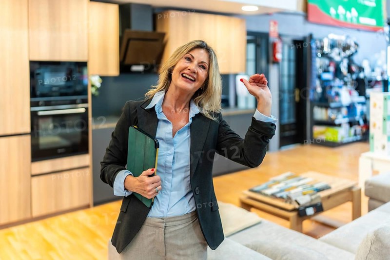 Experienced female advisor standing in a modern showroom, holding a green tablet, smiling and gesturing while discussing home improvement and kitchen design concepts with potential clients