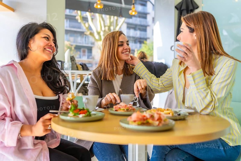 Diverse group of women spending quality time together, happily socializing, drinking coffee and eating brunch while having a lively conversation at a bright modern cafe