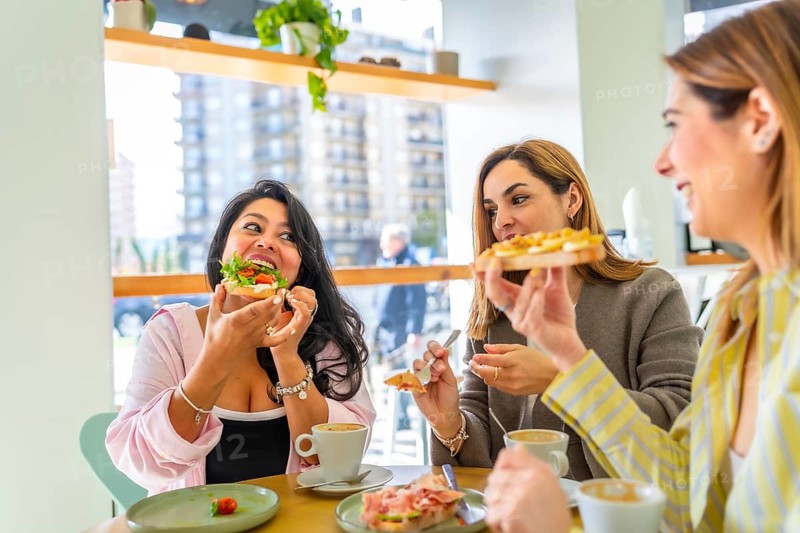 Three diverse female friends sitting at a table in a bright cafe, laughing and eating a fresh healthy brunch, drinking coffee and having a social gathering