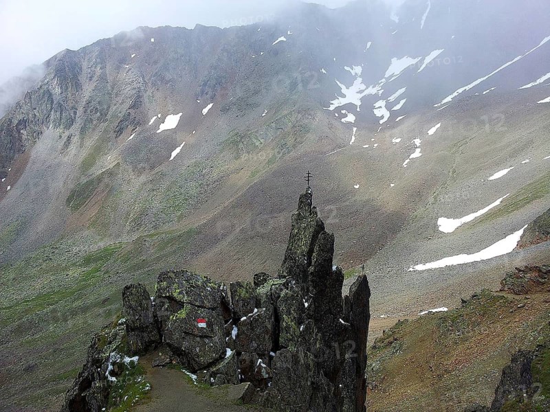 A rocky mountain ridge with red and white markings, signpost, with a summit cross and surrounding fog, E5, hiking in the Alps, crossing the Alps, Austria