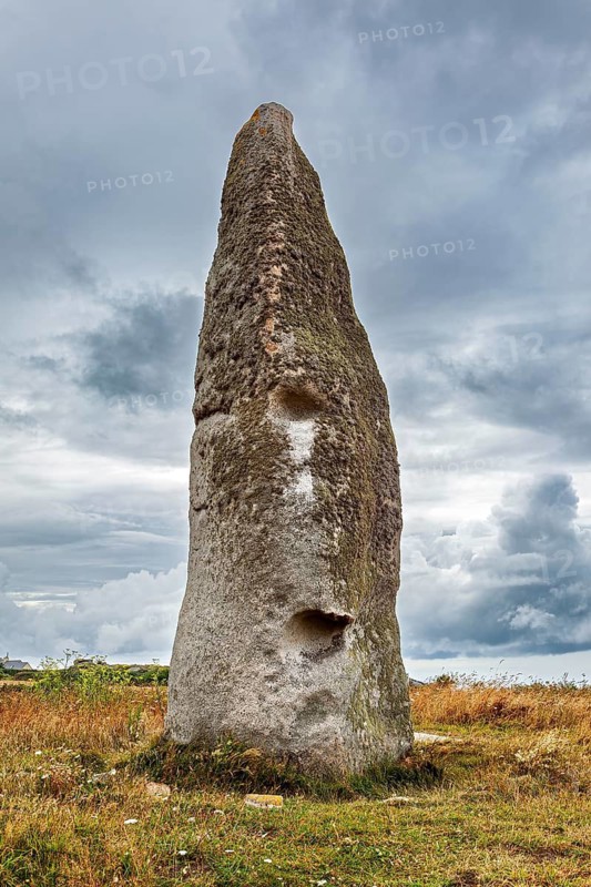 A massive menhir stands in a meadow under a cloudy sky. A monumental impression of the Stone Age, menhir by Cam Louis, a megalithic monument from the Bronze Age in Plouescat, Brittany