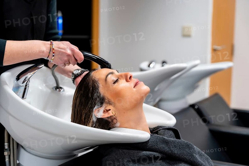 Woman reclining with eyes closed while hairdresser shampoos and conditions her hair at a modern salon basin, enjoying a relaxing professional pampering and hair care treatment