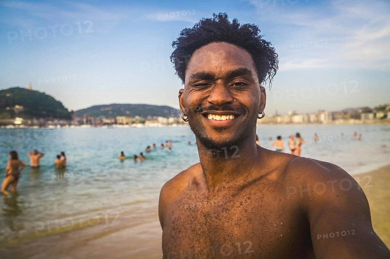 Young adult man winking and smiling into camera during summer vacation, enjoying the sun and refreshing water at a crowded beach with blurred people and coastline in background