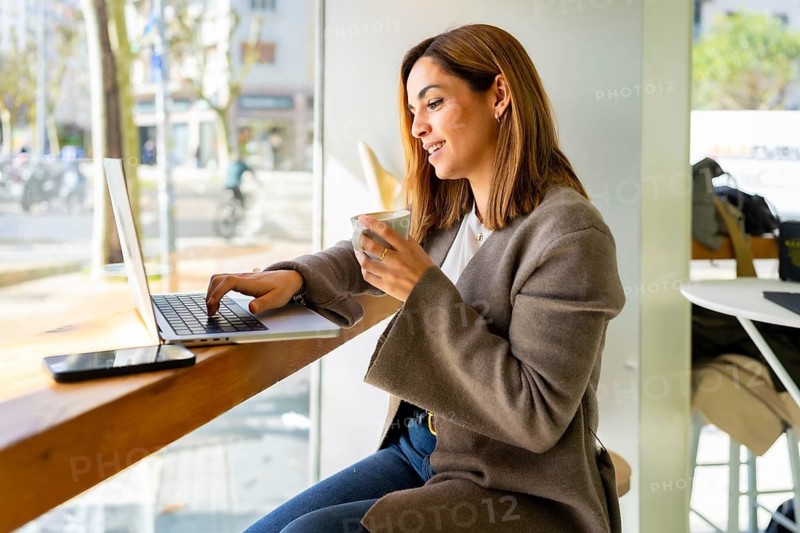 Young smiling woman sitting at a bar table in a cafe, typing on a laptop and holding a cup of coffee, looking out a large window to a busy city street while performing remote work