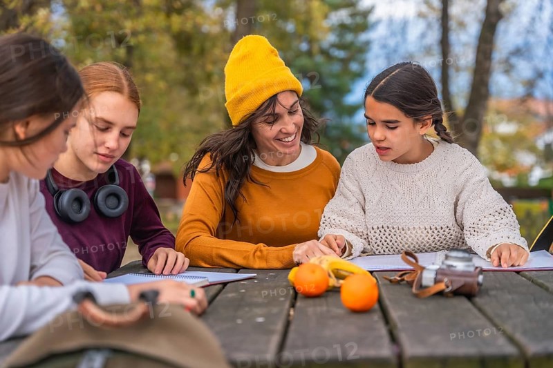 Family enjoying quality time together in a park, mother assisting her two adolescent daughters with their schoolwork and learning on a wooden picnic table during autumn