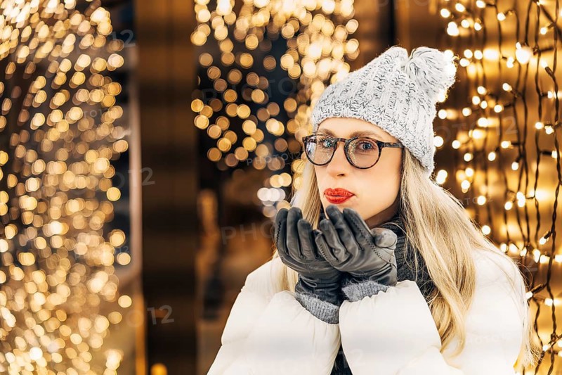 Young woman in knitted hat and down jacket blowing on gloved palms making a wish. Woman wearing white winter coat, knitted hat with pom-pom and gloves stands outdoors surrounded by numerous warm yellow holiday lights. Lights form dense glowing background with pronounced bokeh, creating vivid contrast against night environment. Close framing and bright festive illumination produce visually rich nighttime portrait with strong interplay of warm light, depth and seasonal atmosphere