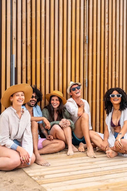 Diverse group of young adults barefoot on a wooden boardwalk, laughing and relaxing together in sunglasses and hats on a sunny summer beach day, carefree and joyful friends