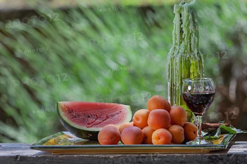 A glass of wine next to a watermelon and apricots against a green background, Provence, France