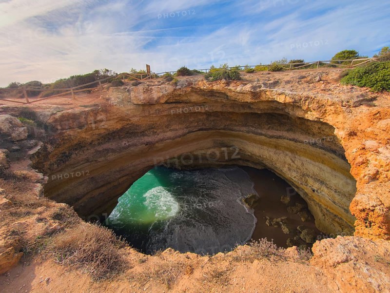 Clifftop view into Benagil Cave in Algarve, Portugal, with a natural cave opening, layered limestone cliffs, Atlantic water, and rugged rock